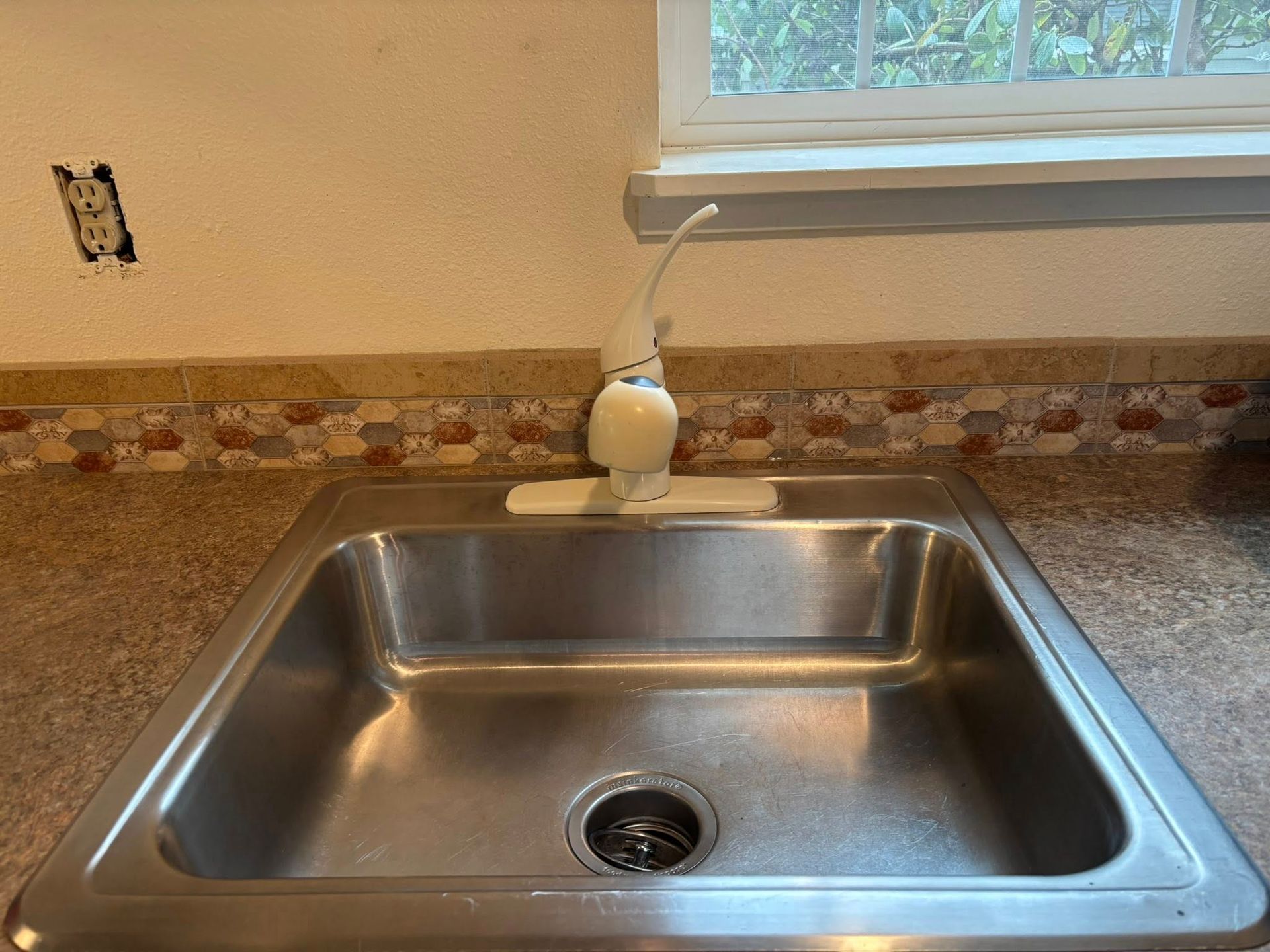 A stainless steel kitchen sink with a white faucet, set into a speckled countertop against a tile backsplash and window.