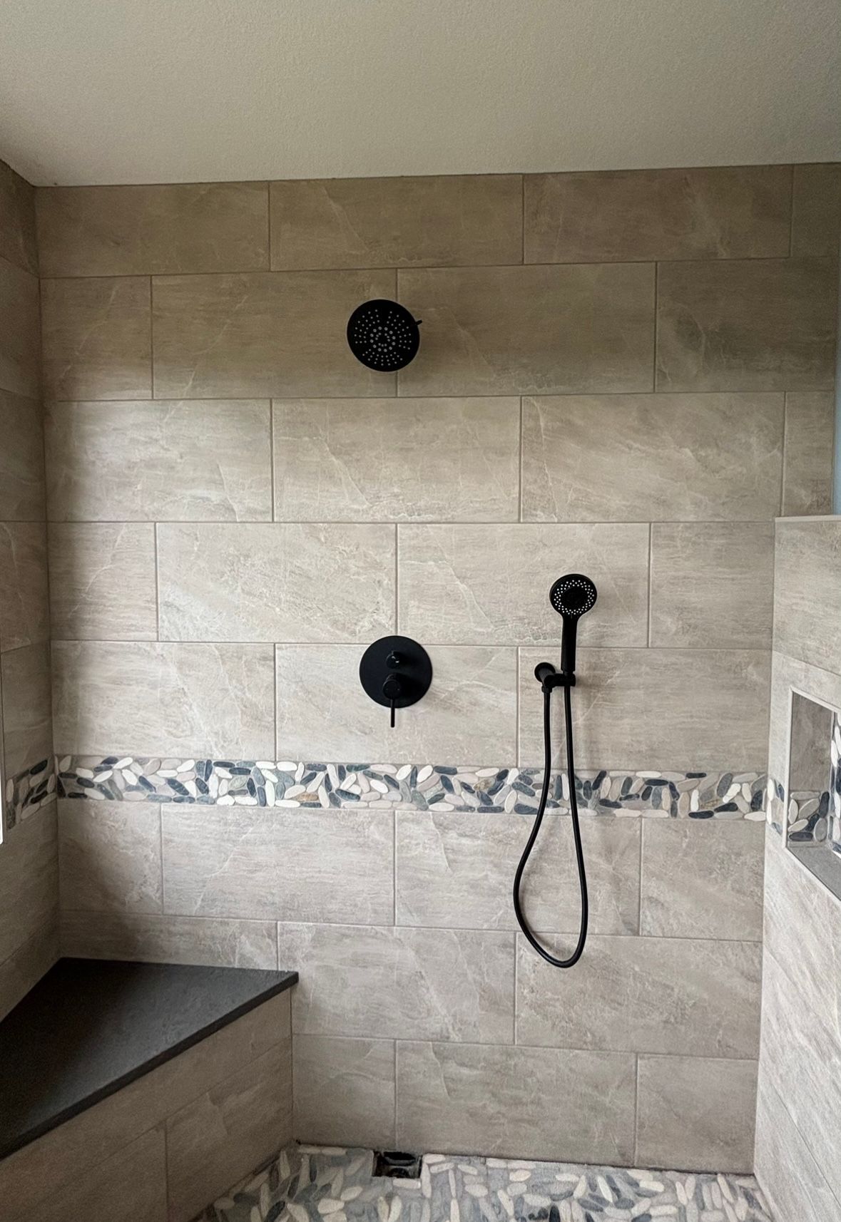 A modern walk-in shower with beige tile walls, a decorative stone border, a corner bench, and black matte fixtures.