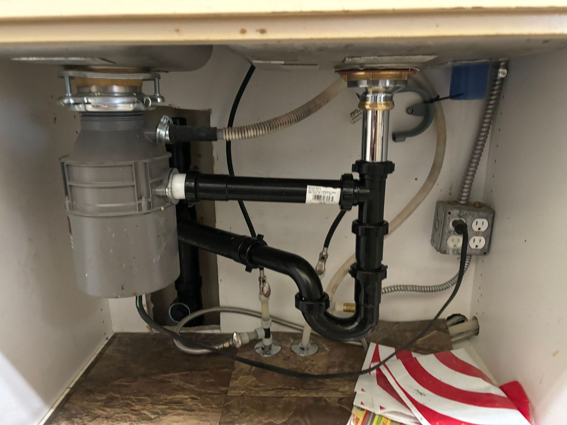 Under-sink view of a garbage disposal unit connected to black PVC pipes, a sink drain, and electrical outlets.