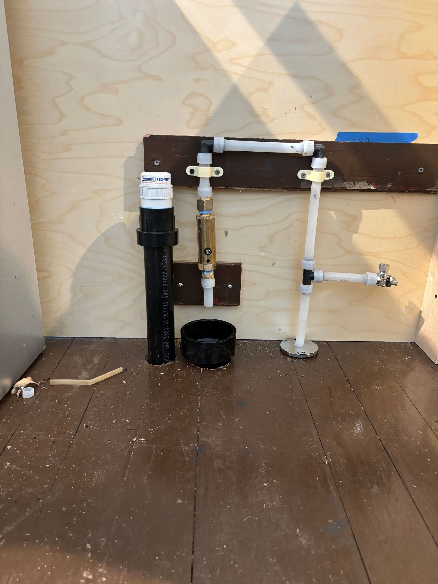 Interior view of a wall showing exposed white PEX water pipes, a brass valve, and a black PVC drain pipe against plywood.