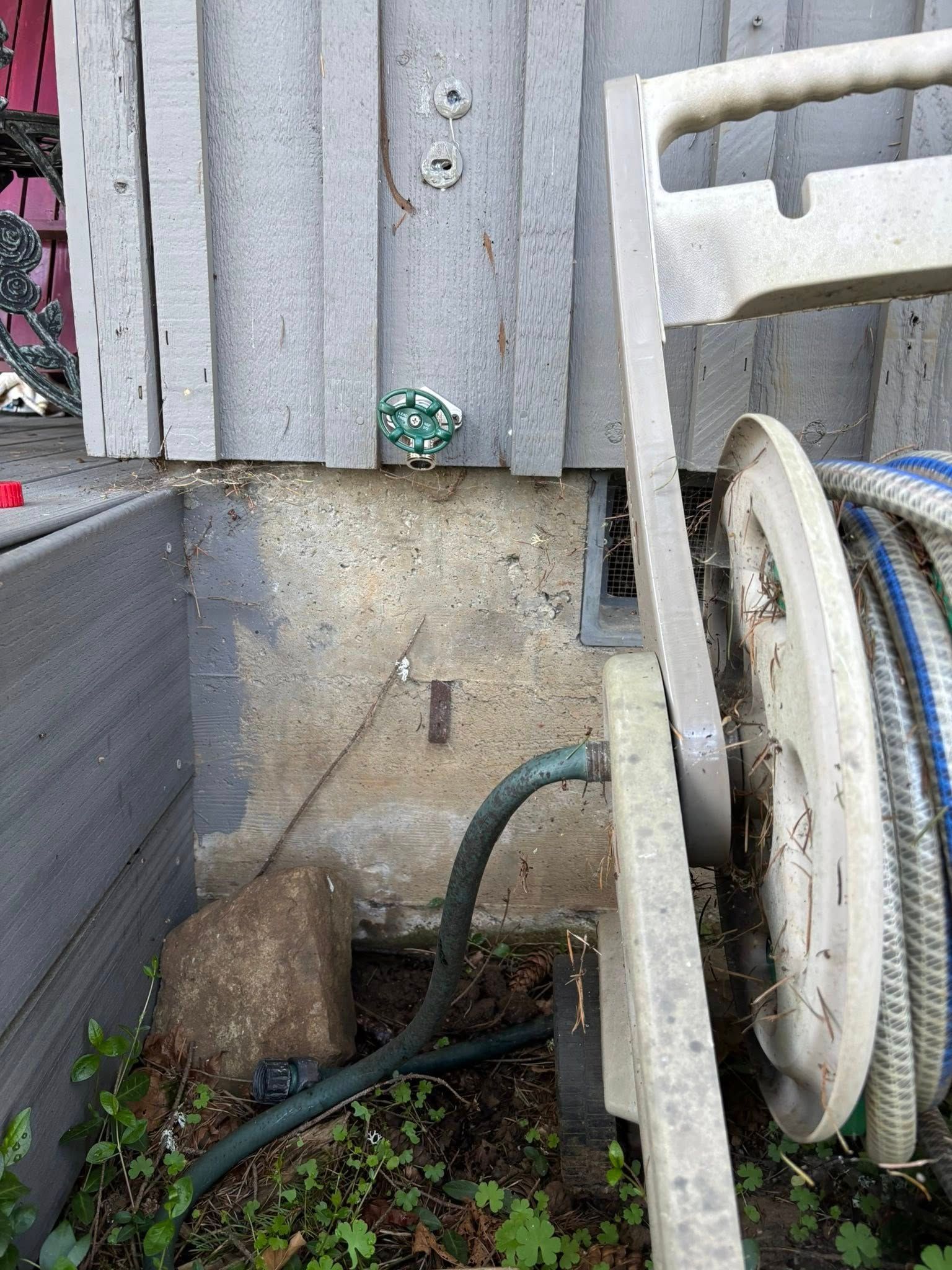 A garden hose on a reel connected to an outdoor faucet on a grey concrete foundation against a wooden-sided building.