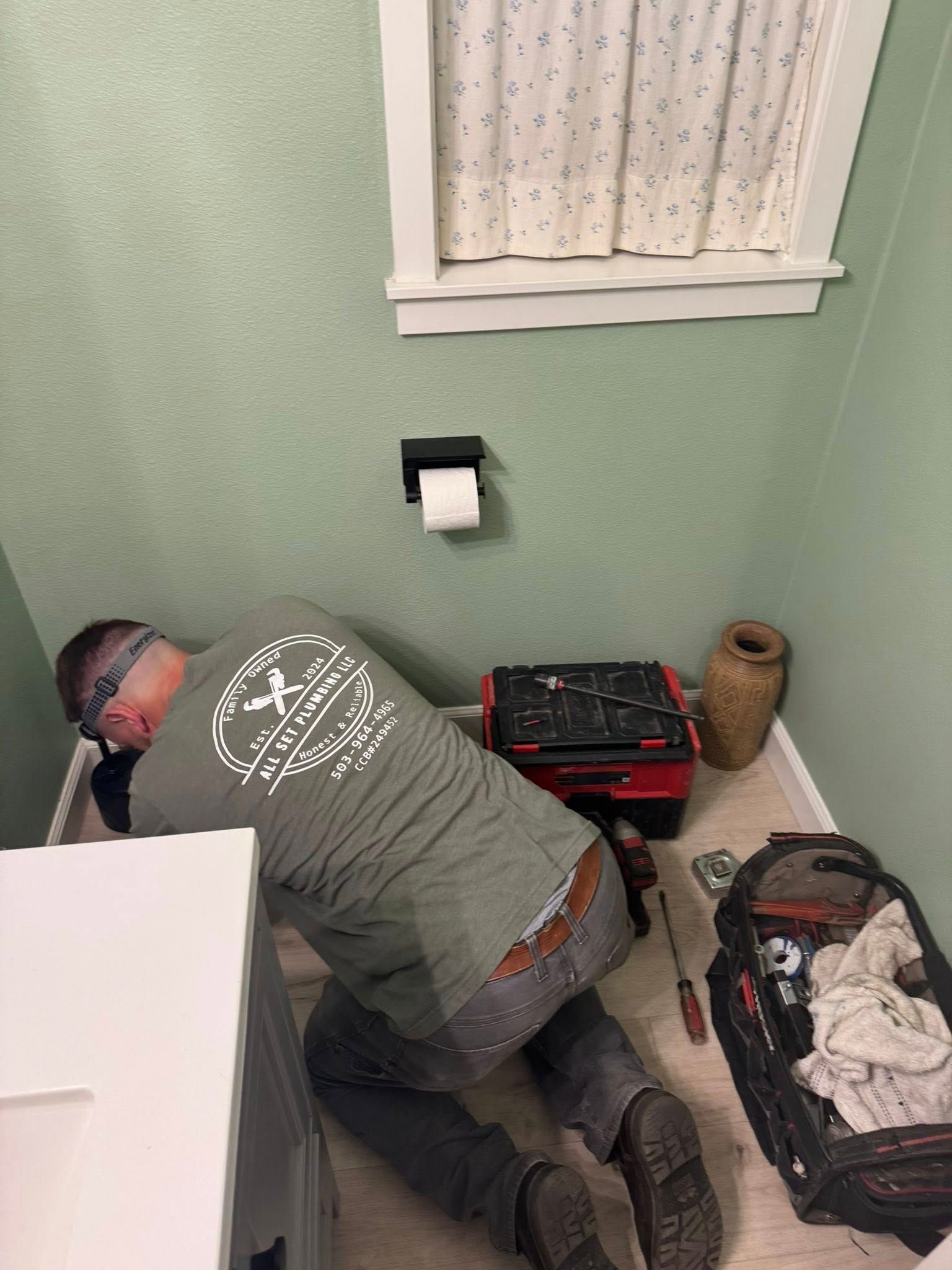 A person wearing a headlamp kneels on a bathroom floor, working on plumbing repairs near a toilet, with tool bags nearby.