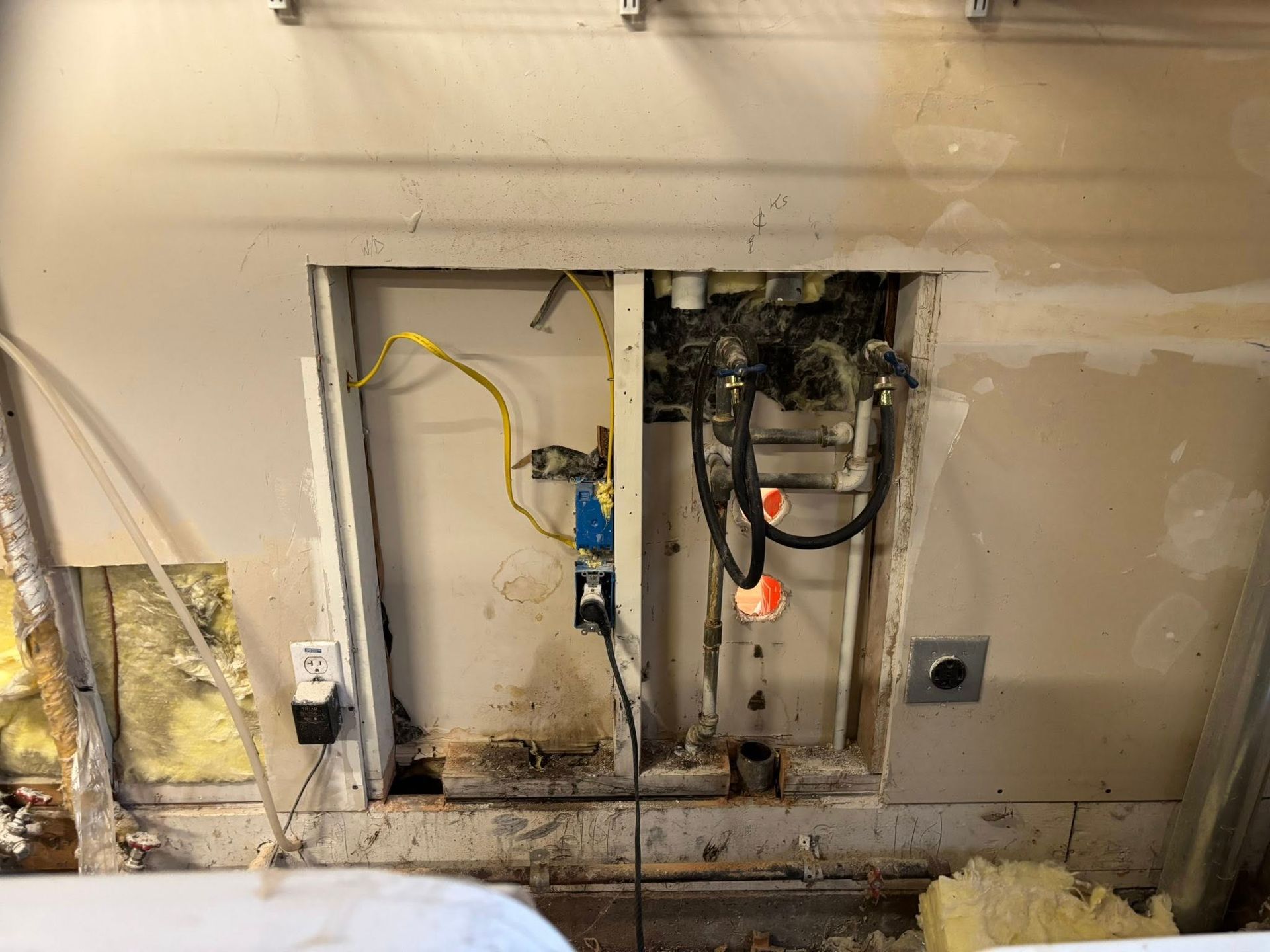 A rectangular hole in a drywall partition revealing exposed electrical wiring, plumbing pipes, and metal wall studs.