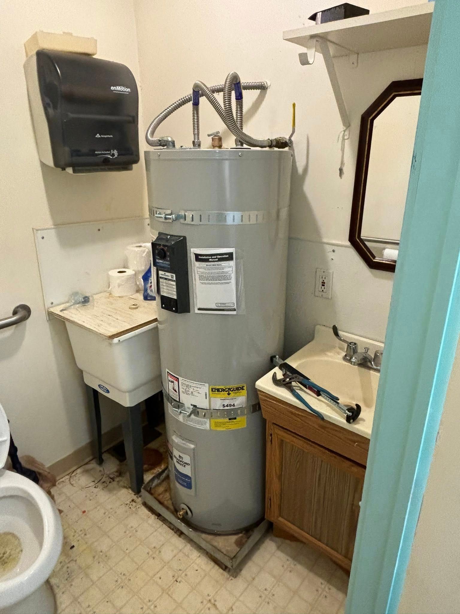 A tall gray water heater sits between a utility sink and a small bathroom vanity with a mirror on the wall above.