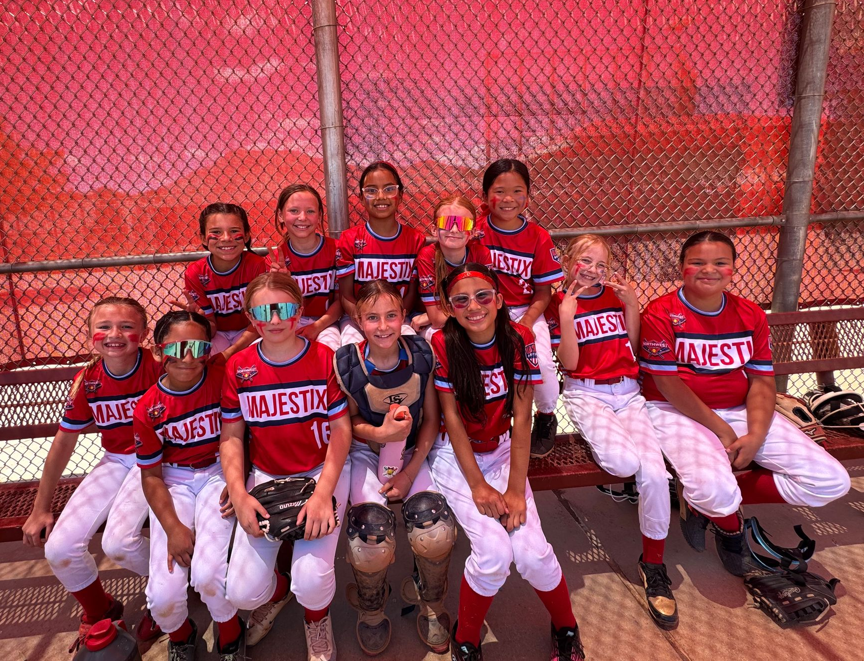 Youth softball team posing in red uniforms on a bench beside a chain-link fence.