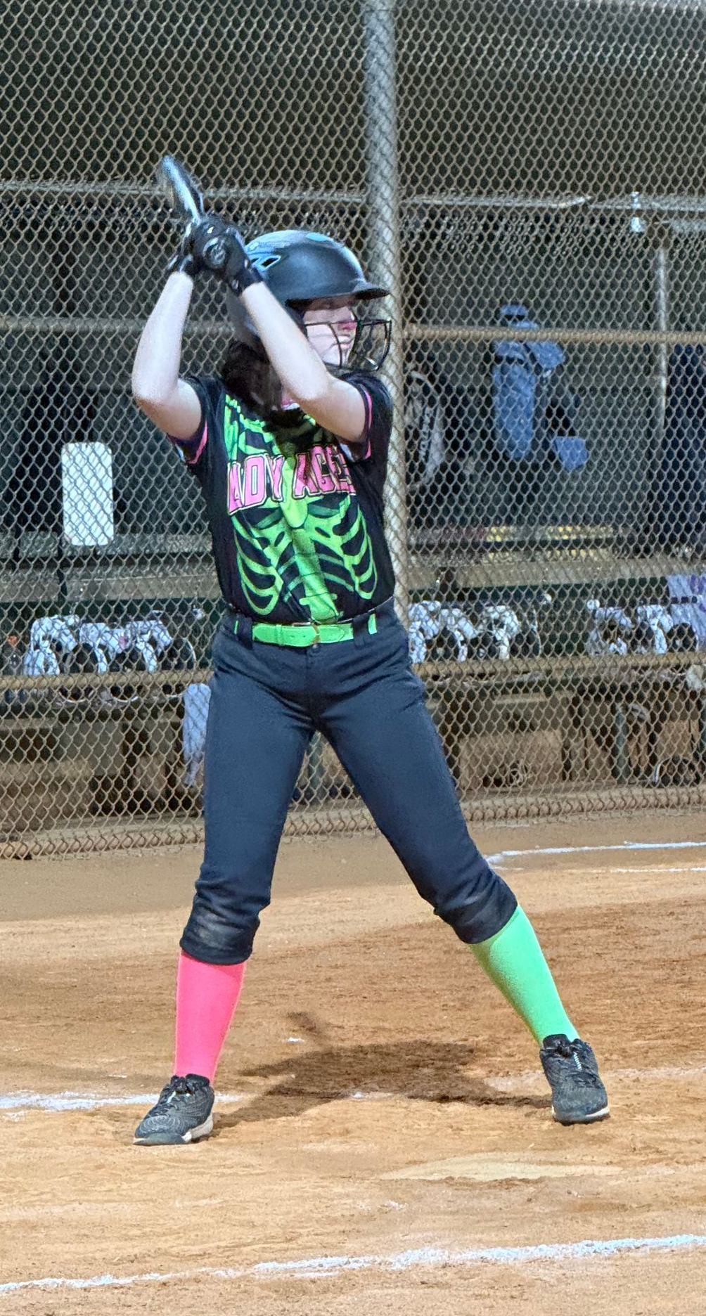 Softball batter in helmet and colorful leggings stands ready at the plate, bat raised in a dirt field.