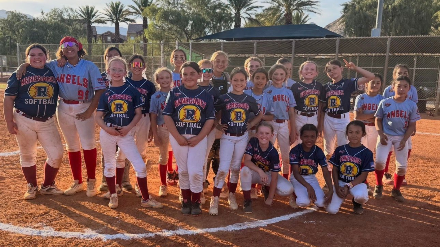 Youth softball team posing on a dirt field in uniform, with orange batters’ boxes and a dugout behind them.
