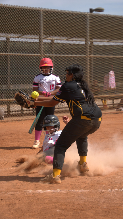 Softball player in black slides into home as catcher waits, dust rising on a sunny field.