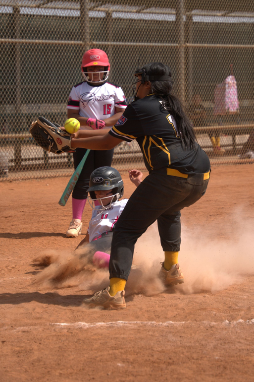 Softball player in black slides into home as catcher waits, dust rising on a sunny field.