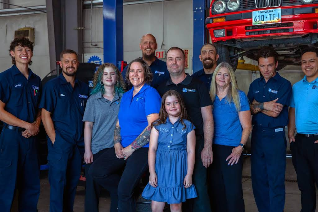 Group of people, mostly in blue shirts, pose in a garage with a car. | Bavarian Auto Haus
