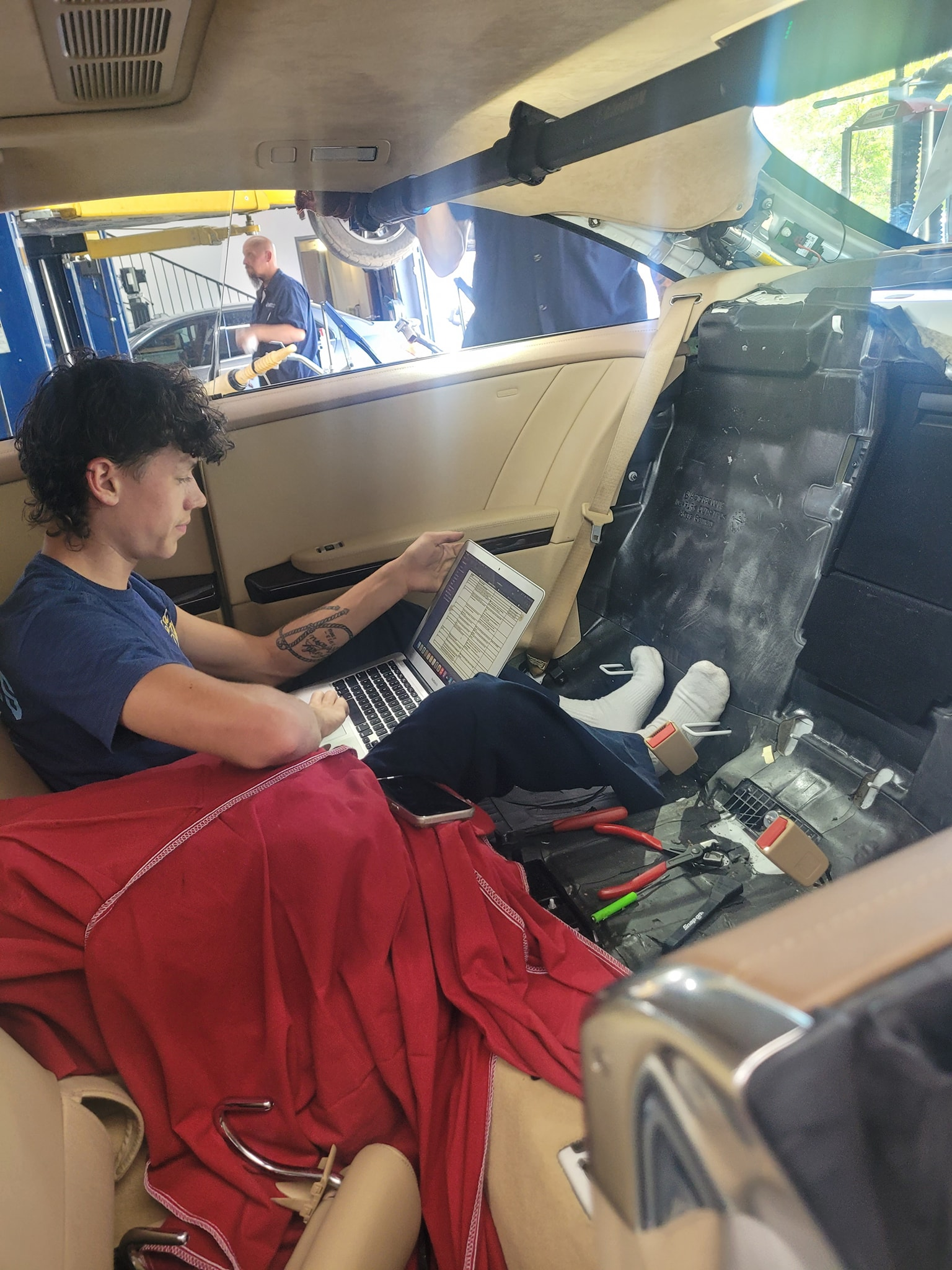 Person using a laptop inside a car, surrounded by tools and red cloth. Another person is in the background. | Bavarian Auto Haus