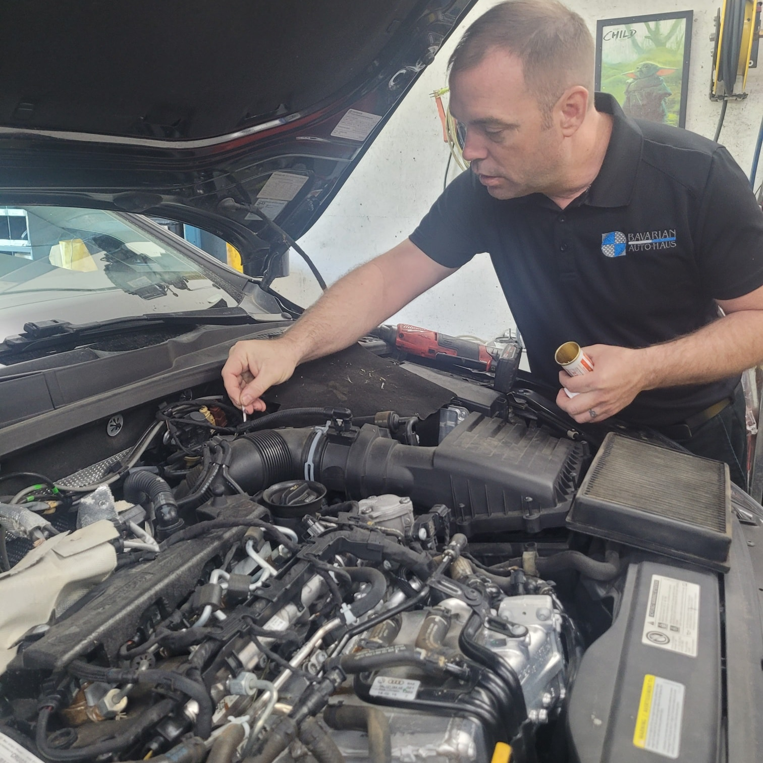 Mechanic working on car engine, inspecting parts. Black shirt, garage setting. | Bavarian Auto Haus