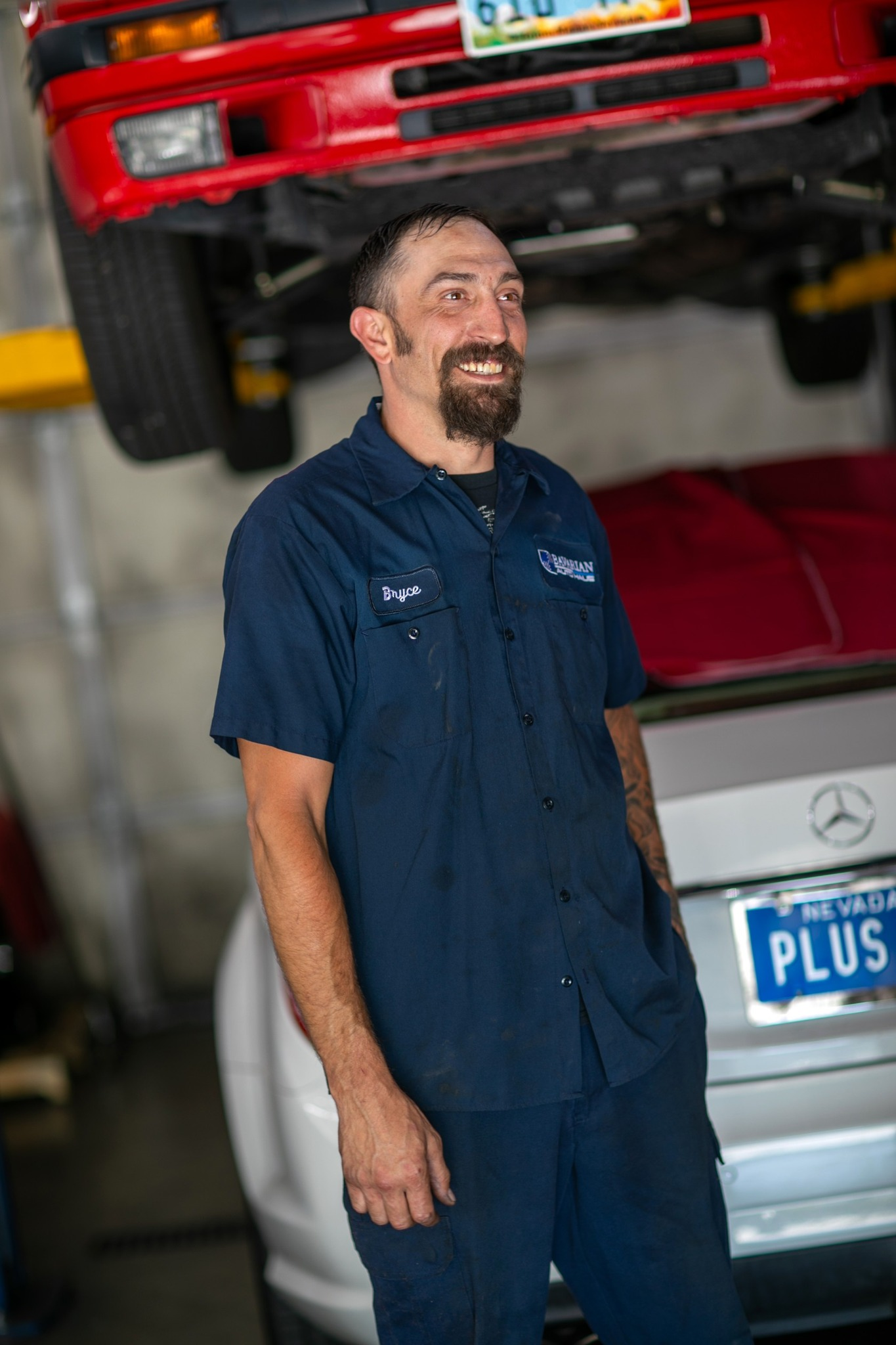 Mechanic in blue uniform smiles, standing in a car repair shop. A red car is above him on a lift. | Bavarian Auto Haus