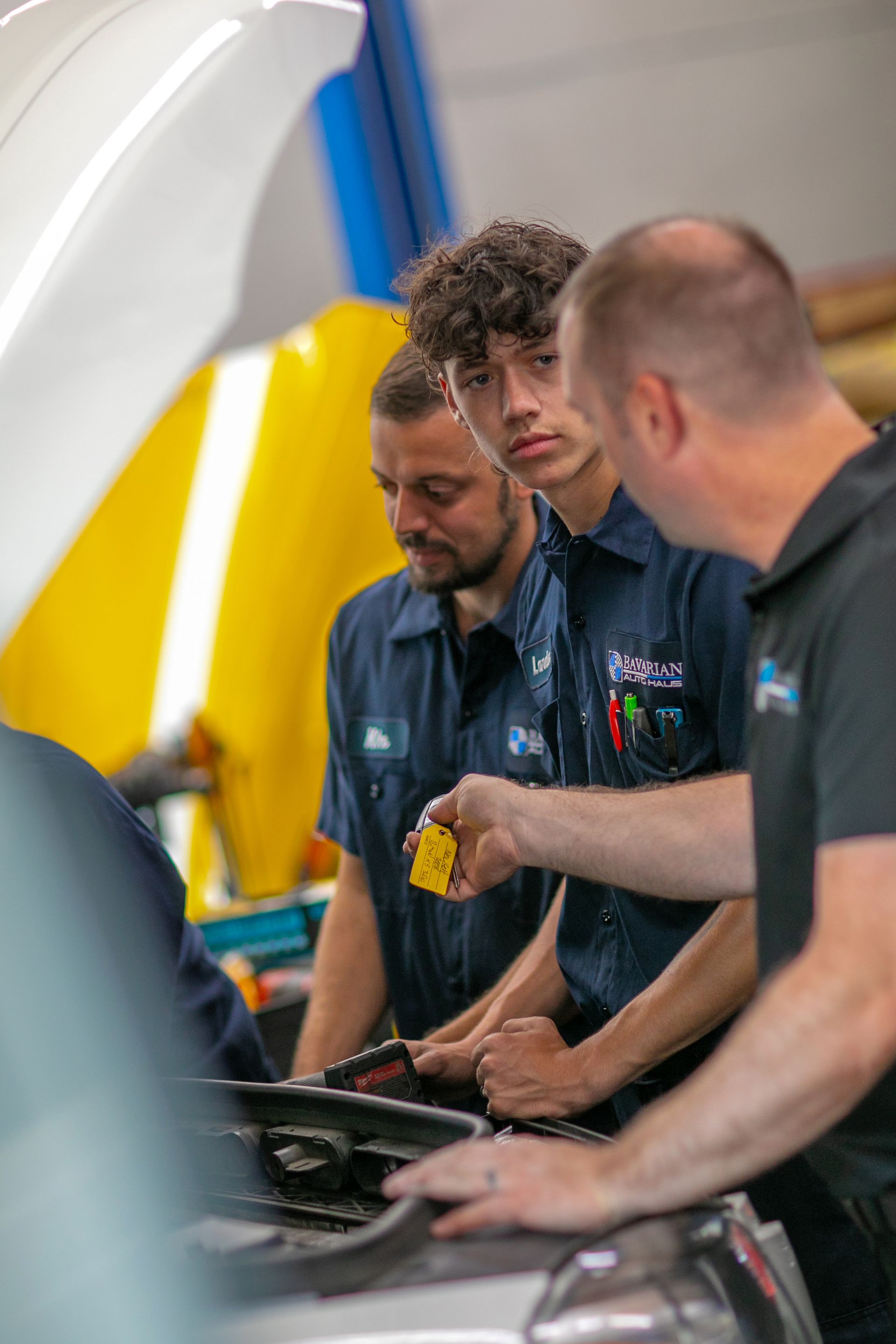 Three mechanics in work uniforms inspect an open car engine, with one pointing to a component while the others watch. | Bavarian Auto Haus