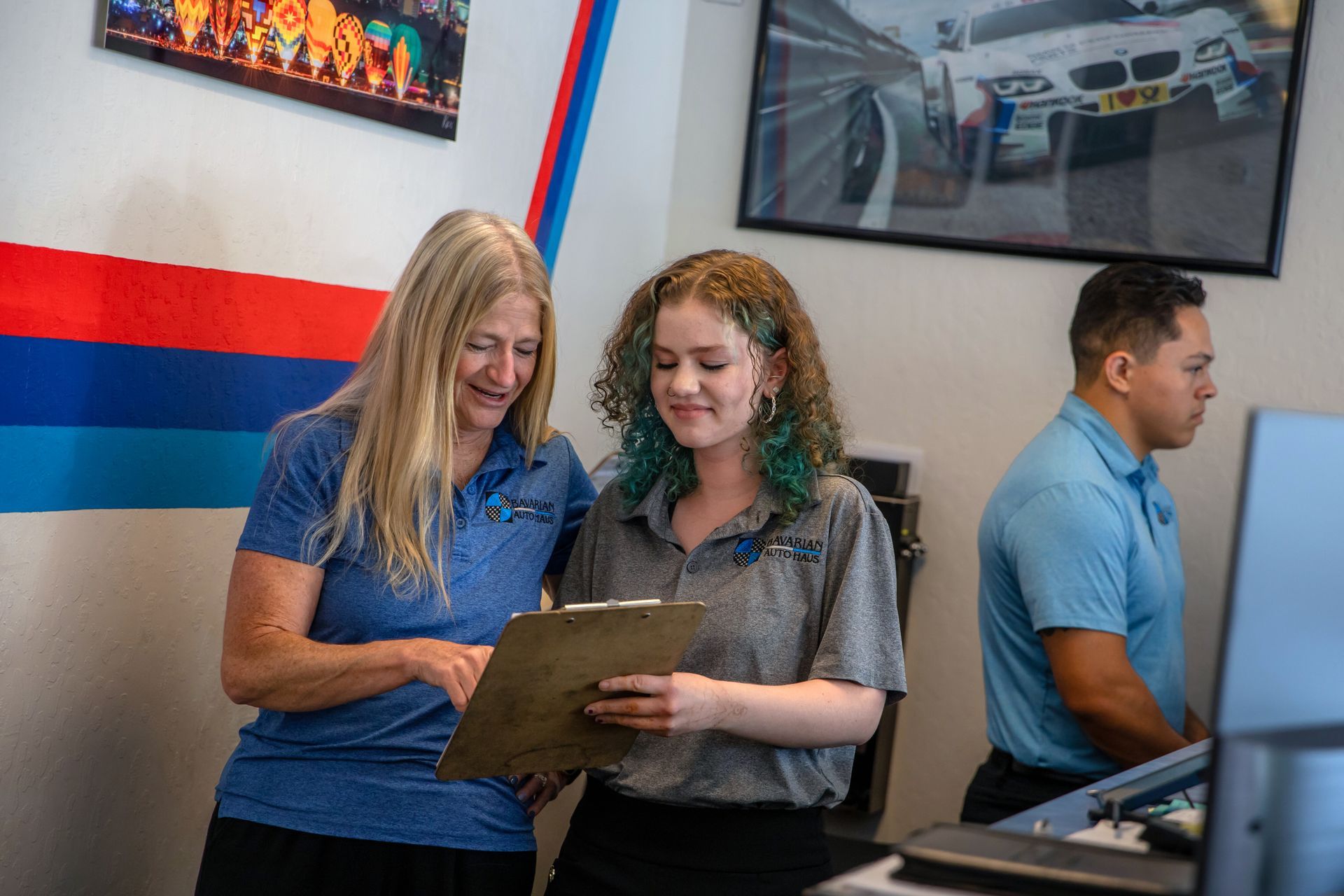 Two people in a professional office setting look down at a clipboard, while a third person works at a computer nearby. | Bavarian Auto Haus