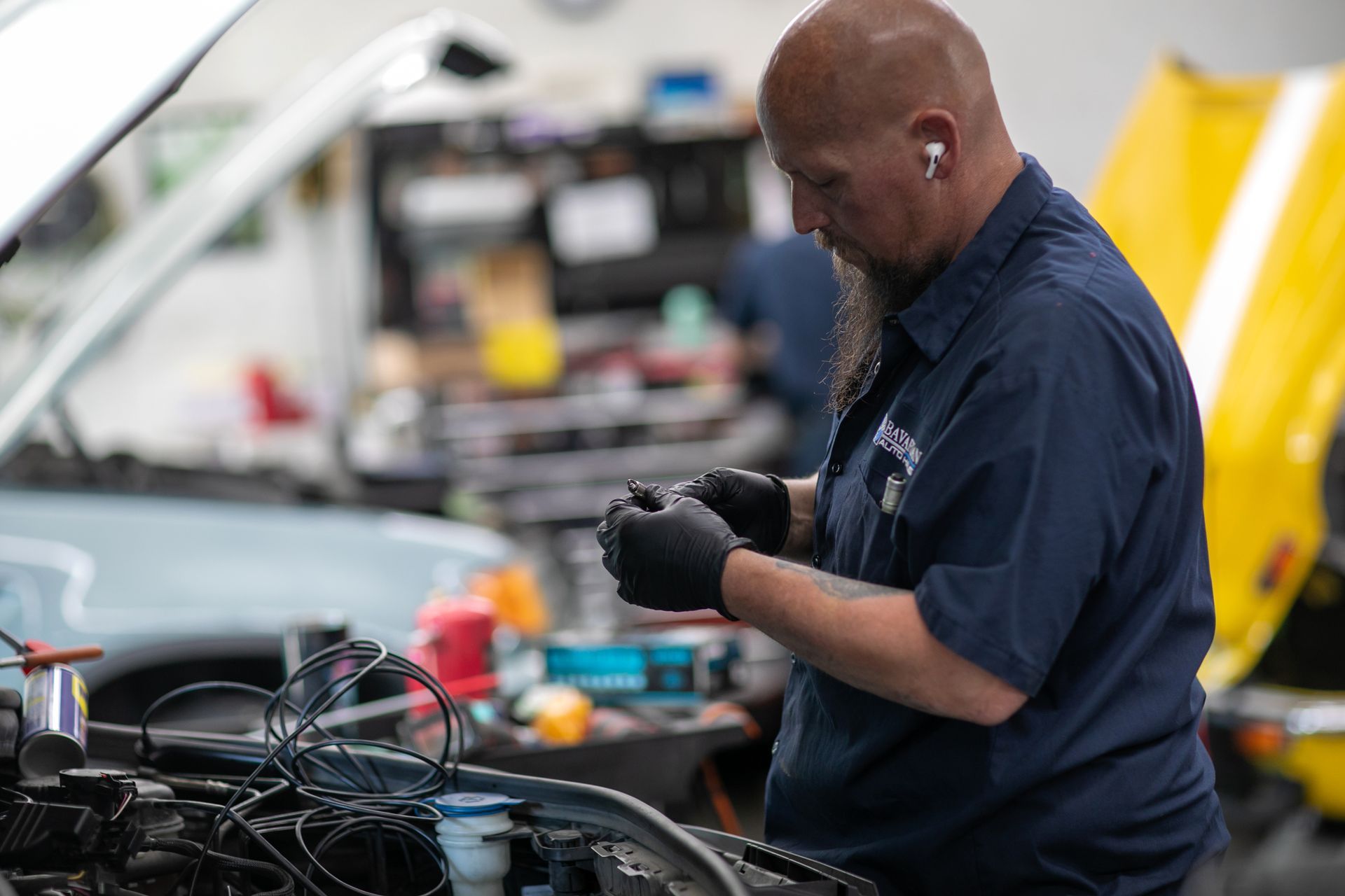 A mechanic wearing black gloves works on an engine inside a brightly lit automotive repair shop. | Bavarian Auto Haus