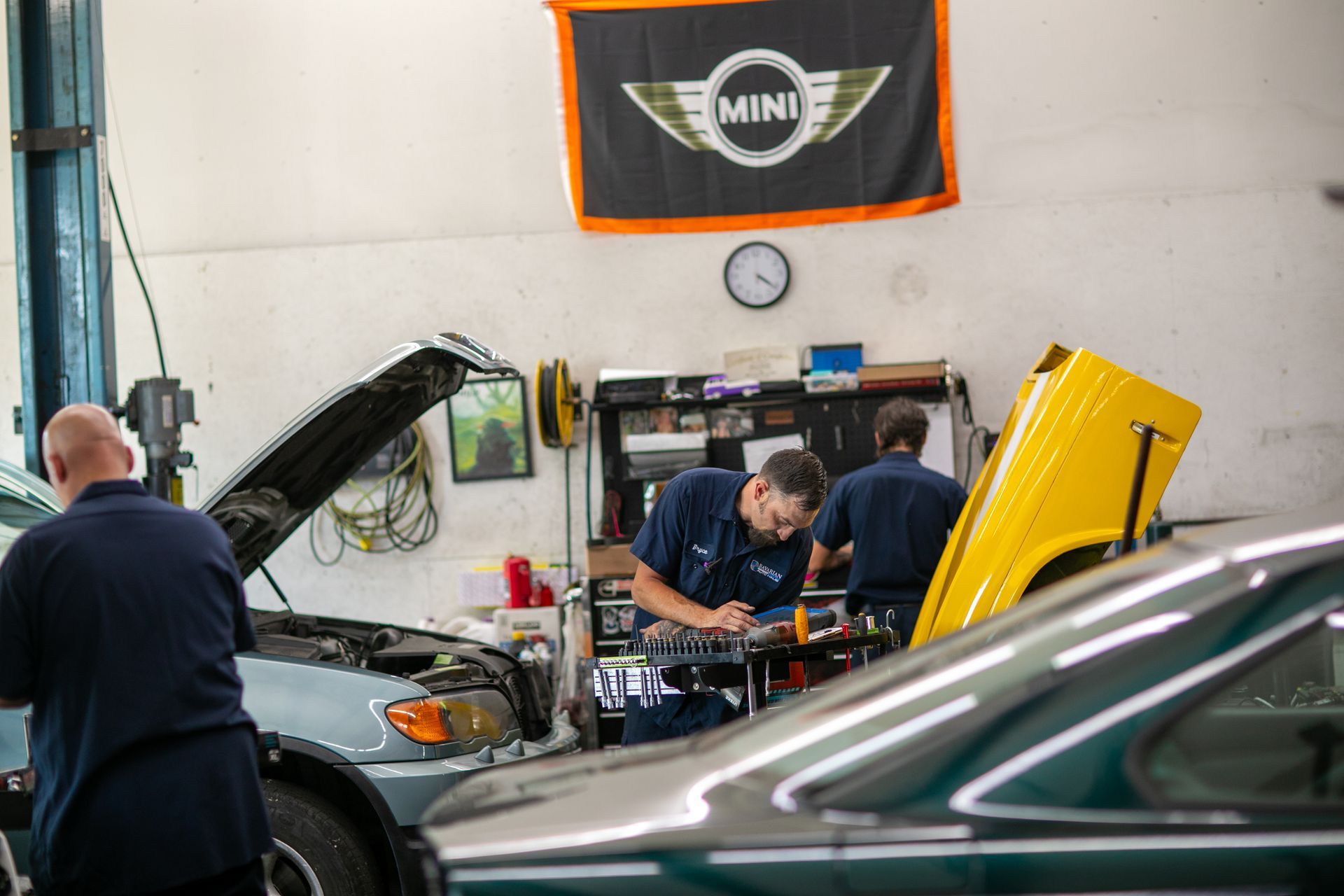 Two mechanics work on vehicles inside a garage with a large MINI flag hanging on the back wall. | Bavarian Auto Haus