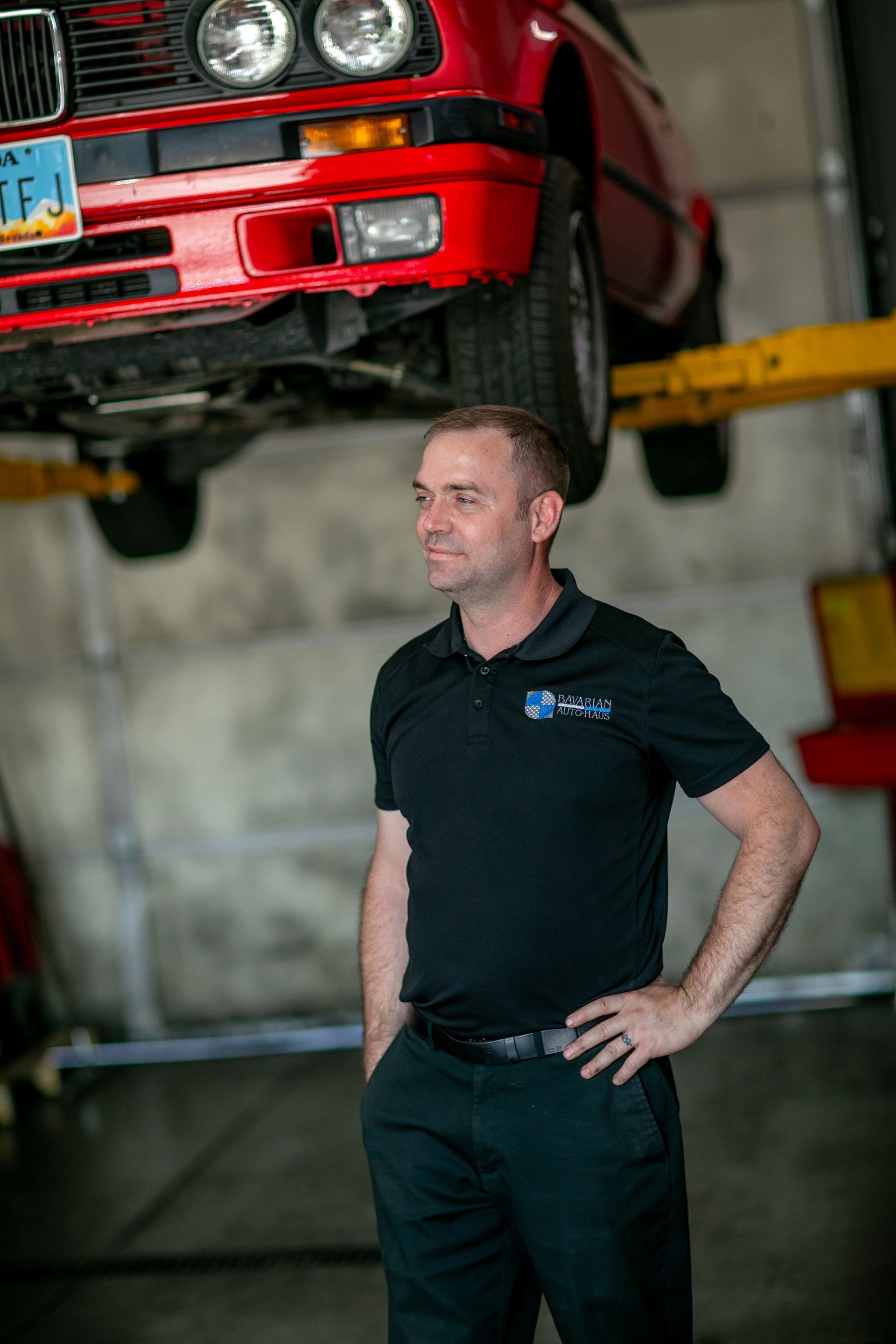 A person in a black polo stands in an auto shop with a red car lifted on a hoist in the background. | Bavarian Auto Haus