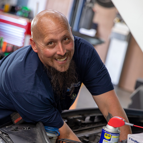 Man working on a car engine, smiling. He has a beard and is wearing a blue shirt. | Bavarian Auto Haus