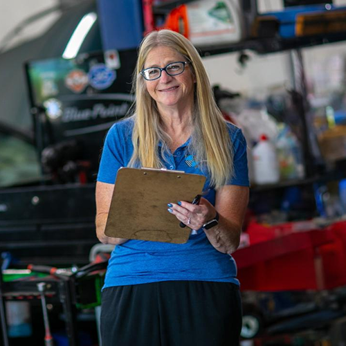 Woman in blue shirt holding clipboard, smiling in auto shop. | Bavarian Auto Haus
