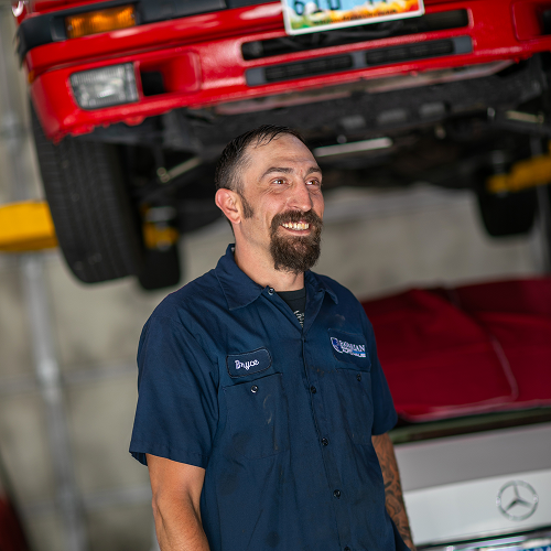 Mechanic smiling in a garage, red car lifted in background. | Bavarian Auto Haus