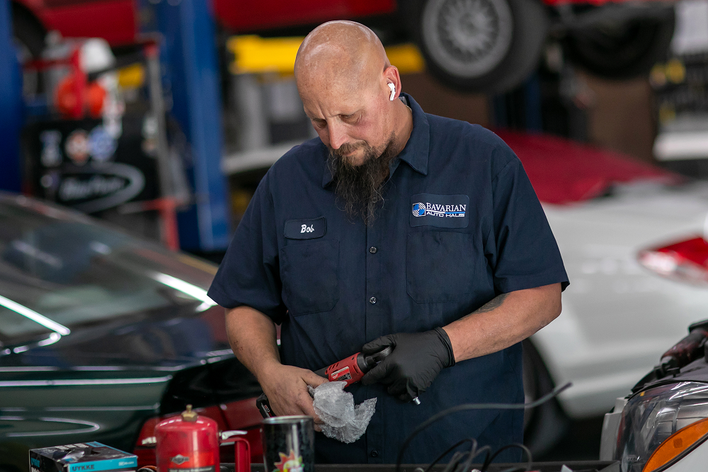Mechanic in blue shirt working on car parts in a garage, using a tool. | Bavarian Auto Haus