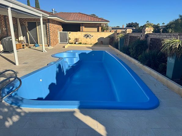 Blue swimming pool beside a brick house with patio and yard, viewed on a sunny day