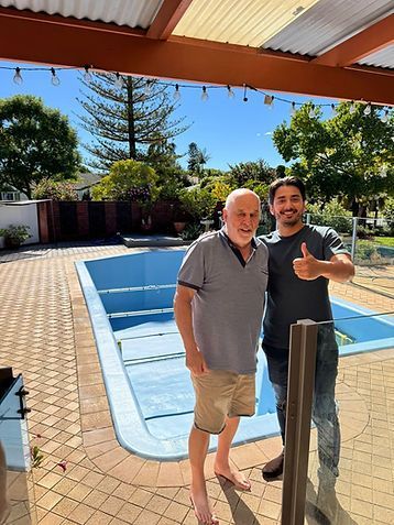 Two men standing by a backyard swimming pool under a covered patio, one giving a thumbs-up
