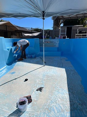 Person painting a backyard pool bright blue under a sun umbrella.