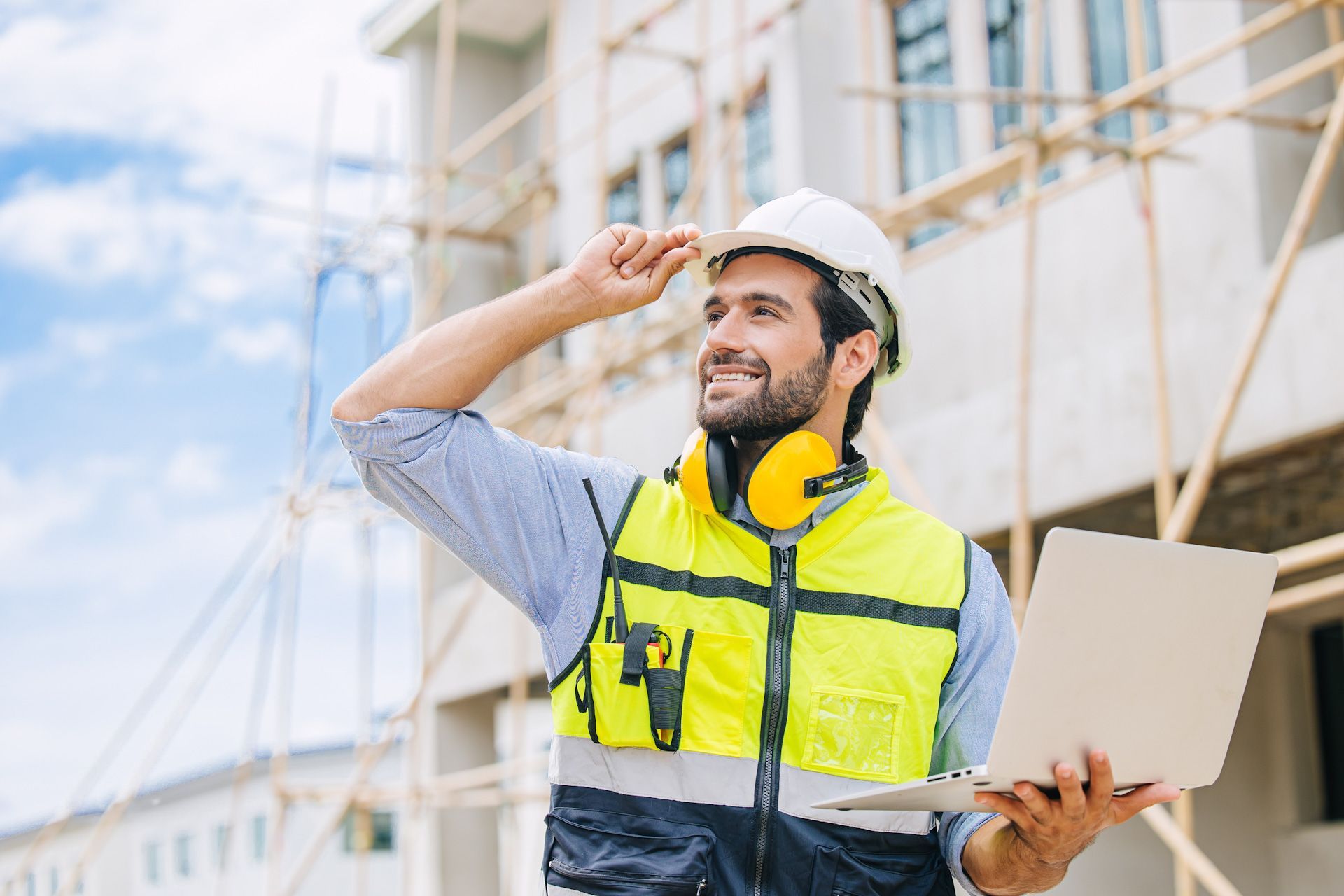 A construction worker is holding a laptop computer in front of a building under construction.