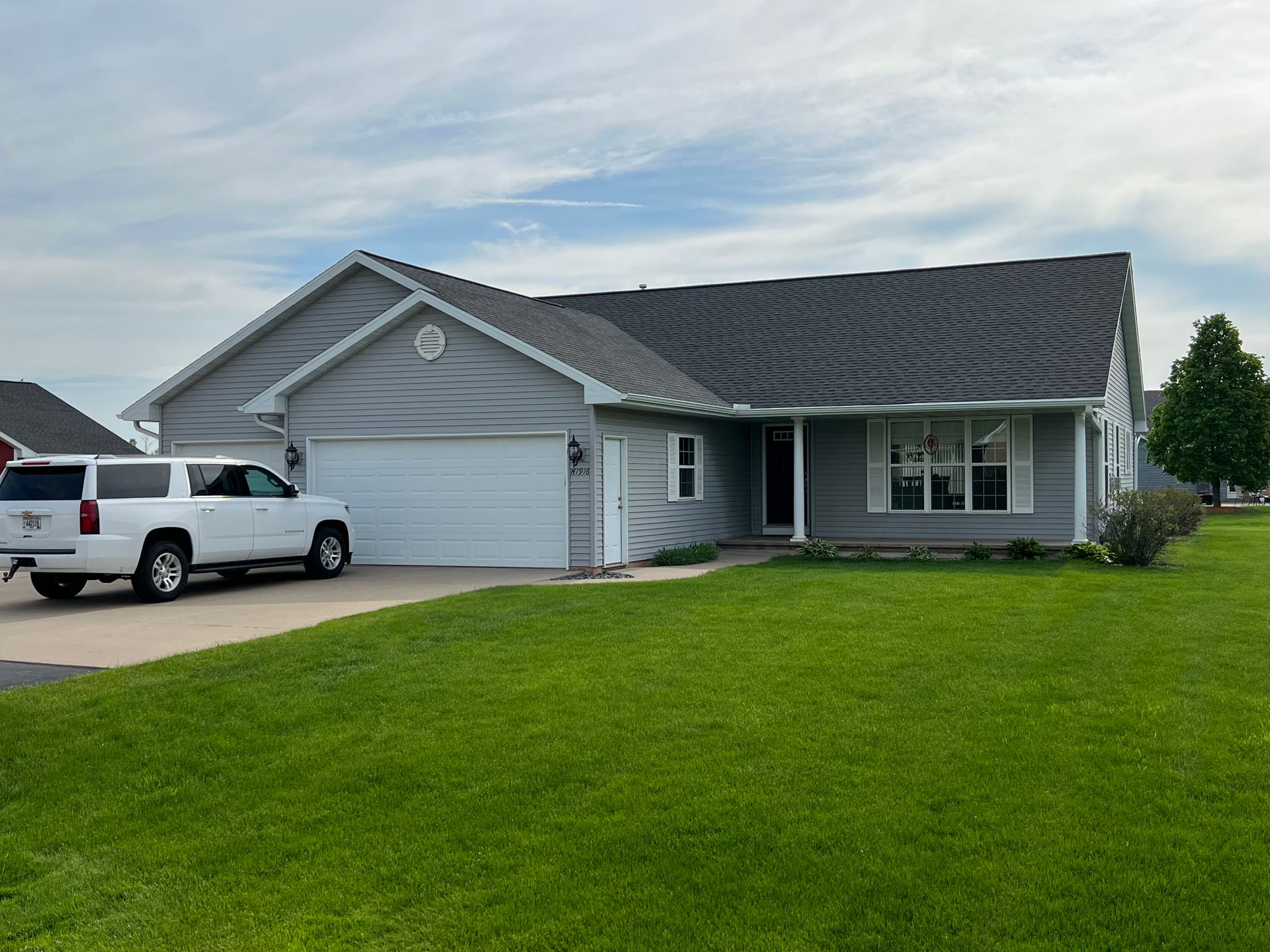 A grey, single-story suburban house with a two-car garage and a white SUV parked in the driveway on a sunny day.