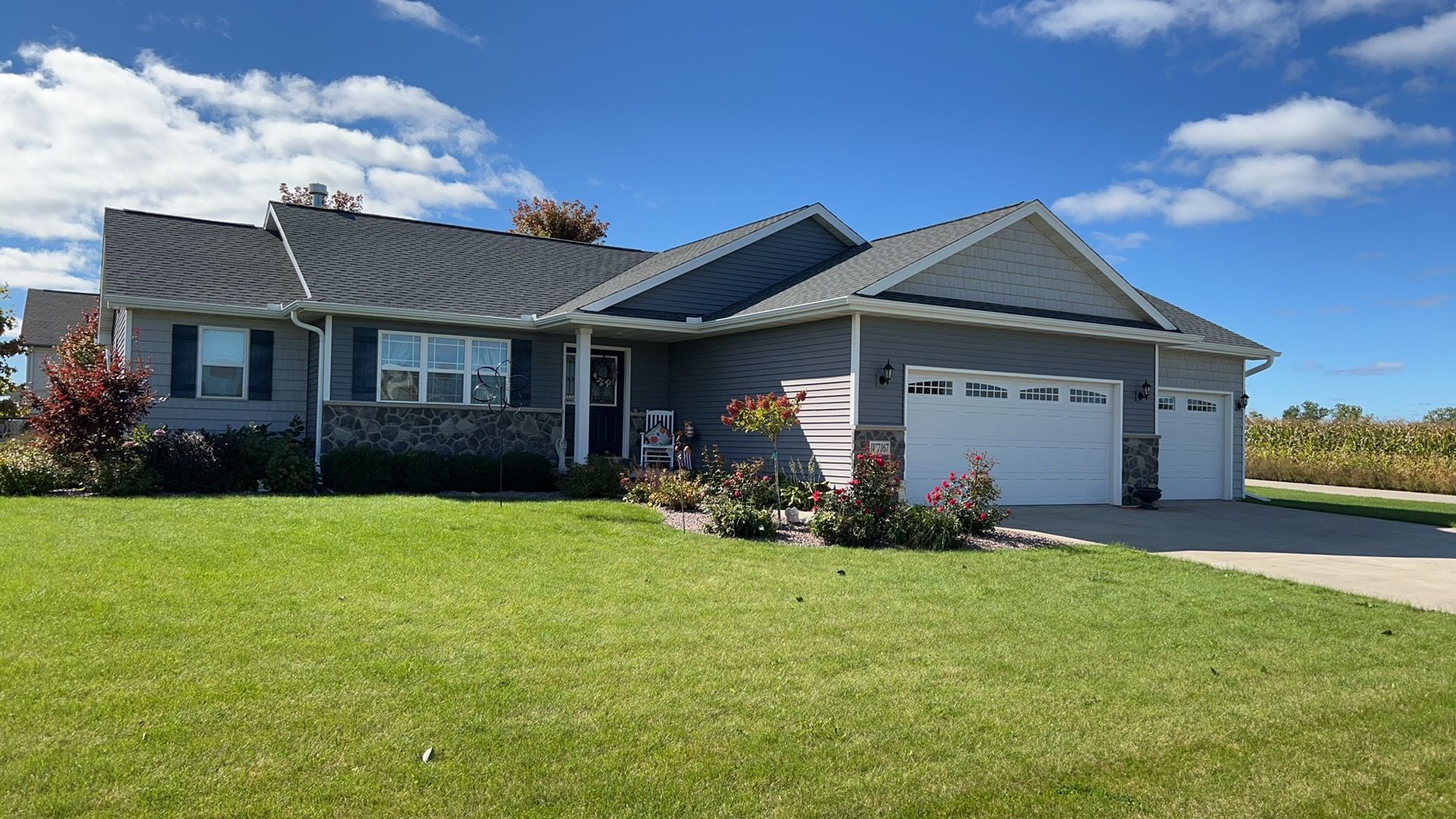 A grey, one-story ranch-style house with a multi-car garage, stone facade accents, and a large lawn under a blue sky.