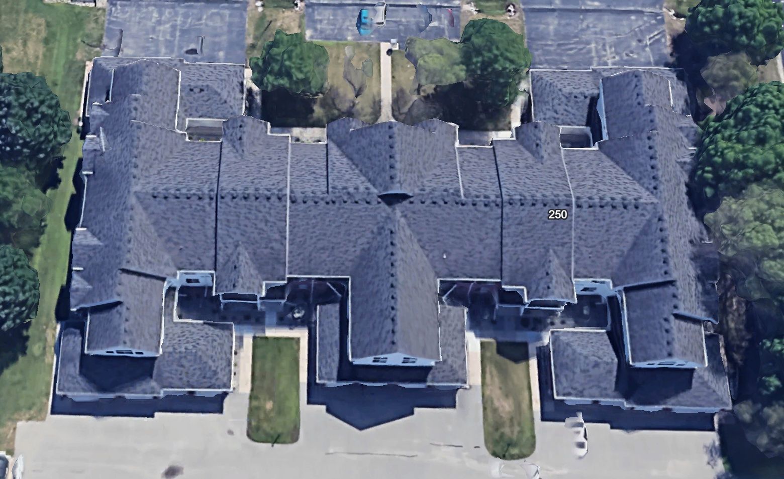 An aerial view of a multi-unit residential building with a dark grey shingled roof surrounded by parking lots and trees.