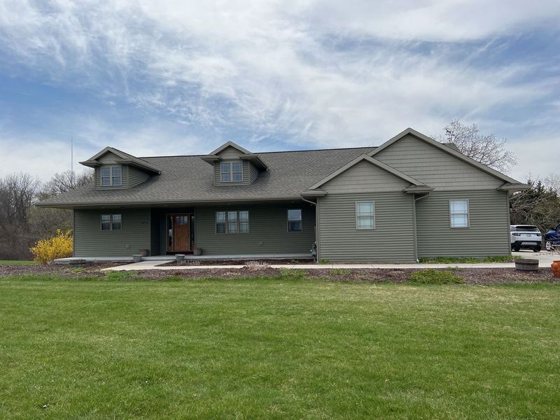 A single-story, olive-green house with a dark shingled roof, two dormer windows, and a paved front walkway.
