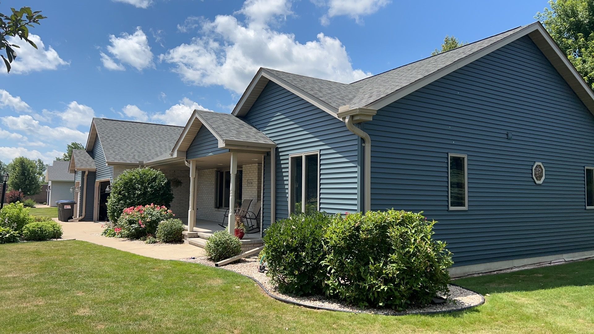 A blue-sided, one-story house with a grey roof and manicured bushes in the front yard under a bright, sunny blue sky.