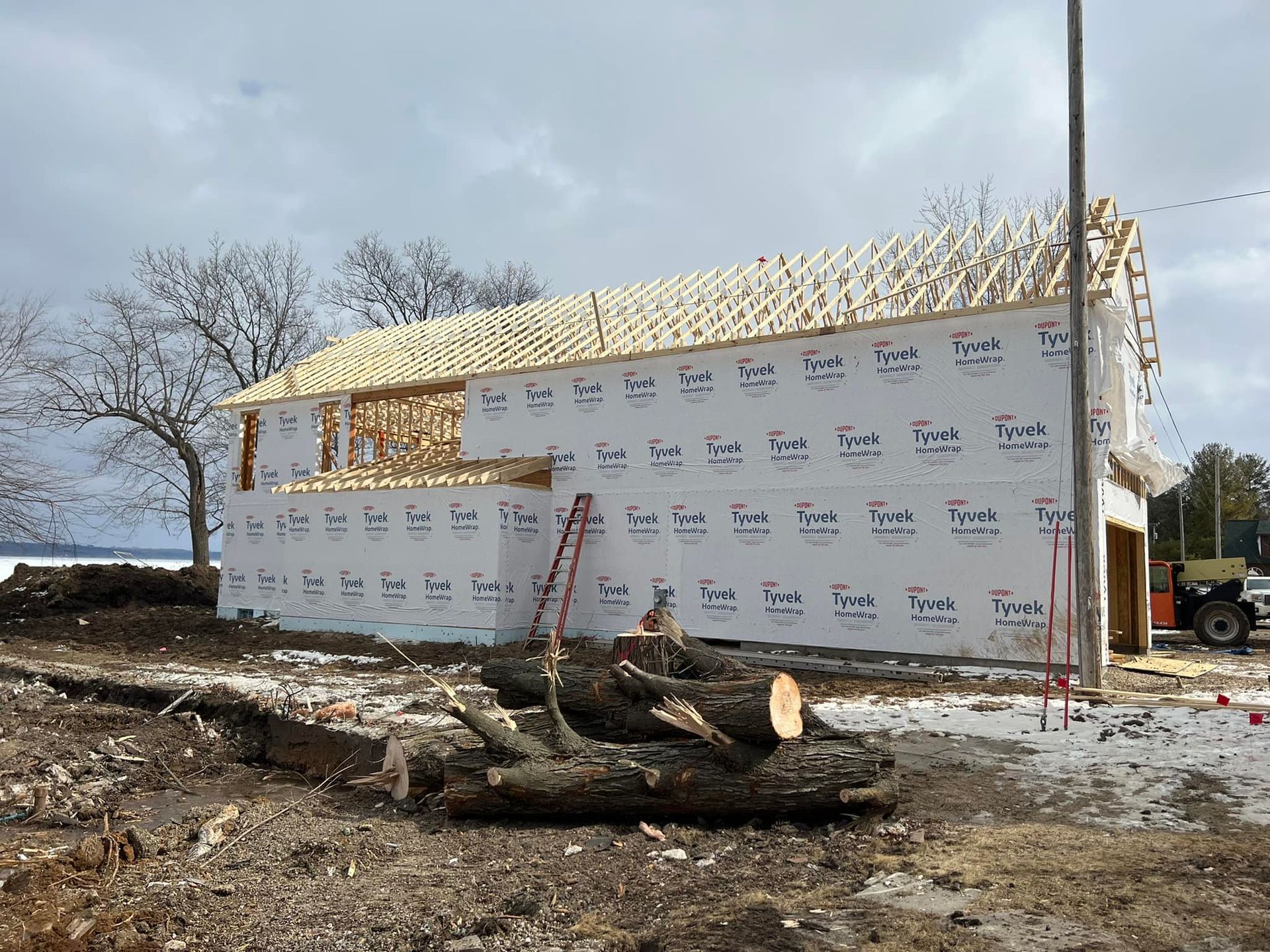 A house is being built in a field with a pile of logs in front of it.