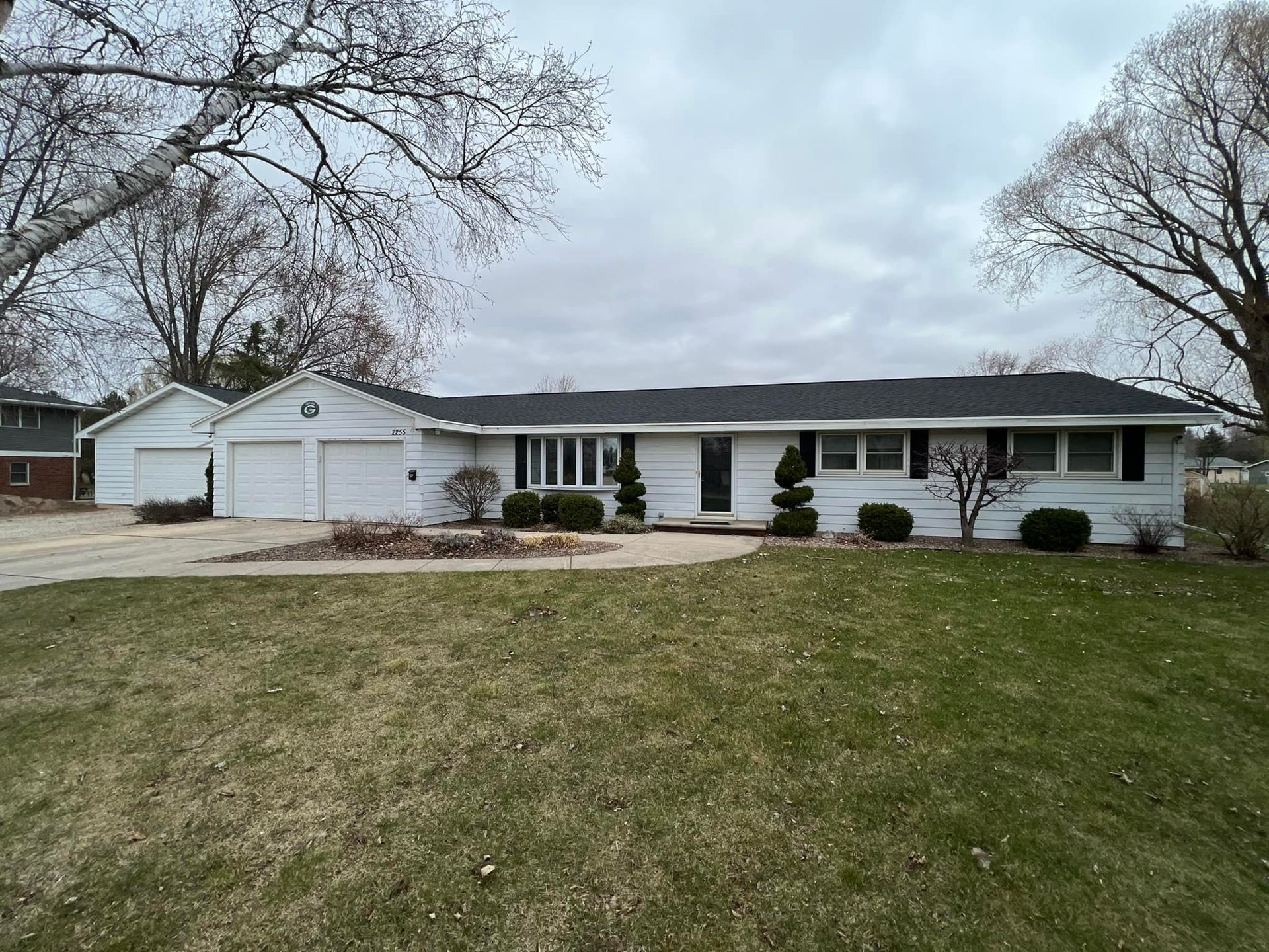 A white, single-story ranch-style house with a two-car garage, gray roof, and a large front lawn on a cloudy day.