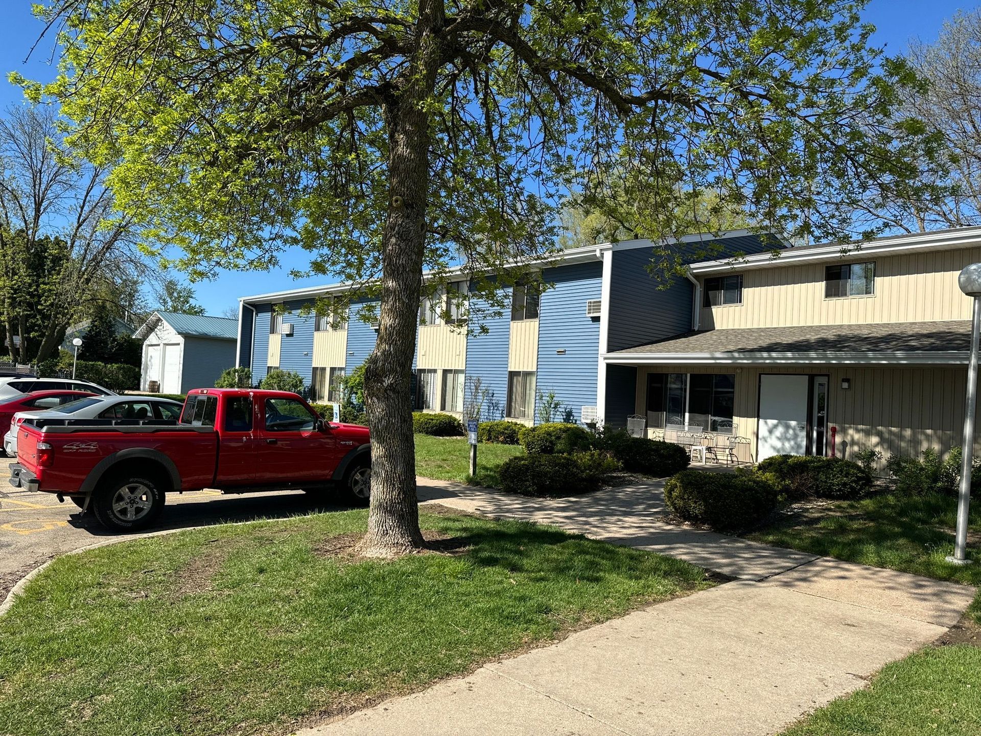 A multi-story apartment building with blue and tan siding next to a parked red pickup truck on a sunny day.
