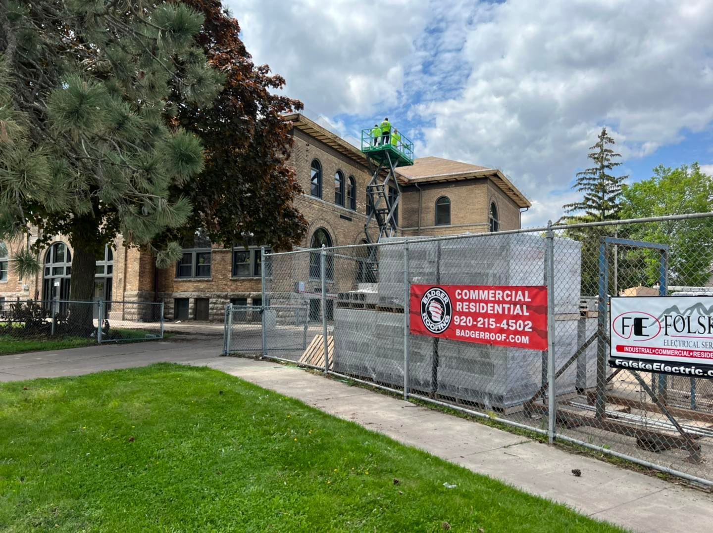 A brick building under renovation with construction materials behind a chain-link fence and a small lift on the roof.