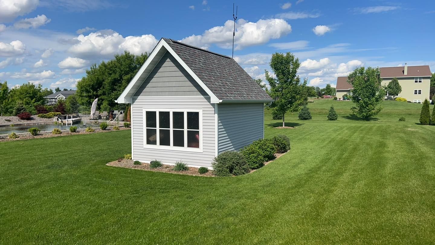 A small, light-gray shed with a dark shingled roof and a large window sits in the middle of a wide, sunny green lawn.