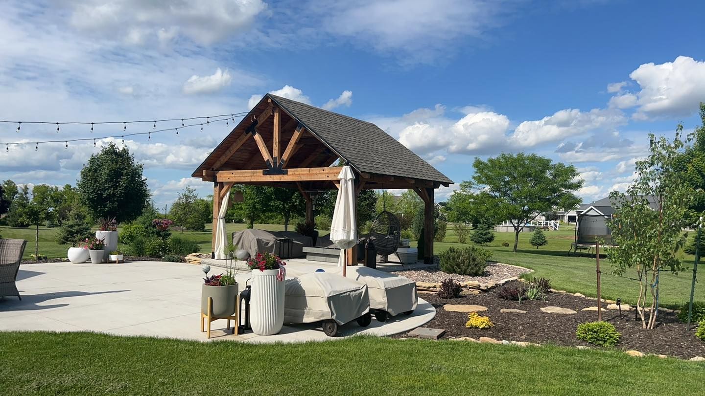 A wooden pavilion with a peaked roof over a patio, featuring outdoor furniture with covers on a sunny, grassy backyard.