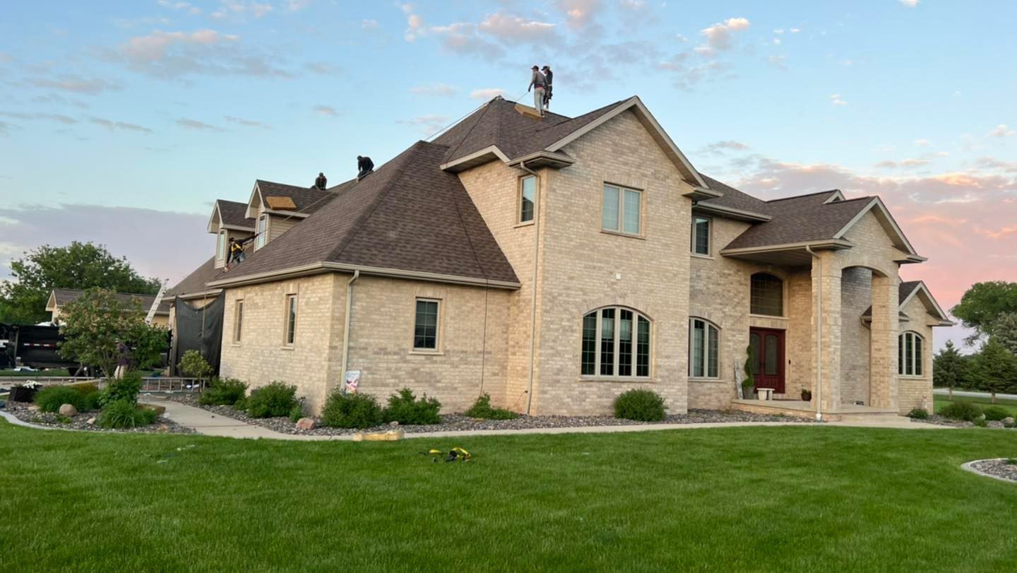 A multi-story beige stone house with a dark shingled roof stands on a green lawn under a sunset sky.
