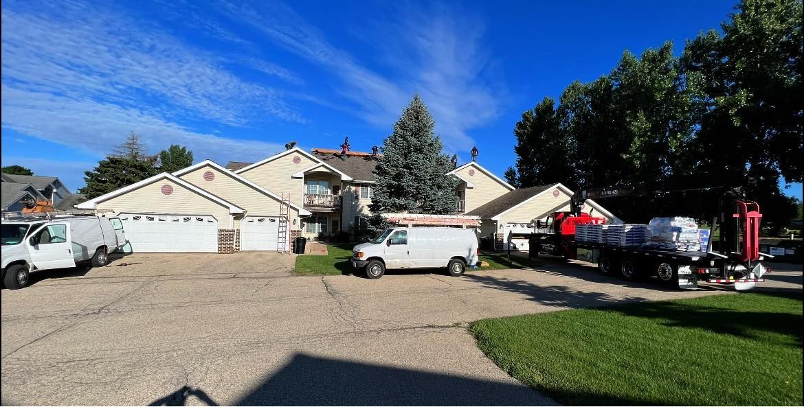 Service vans and a trailer loaded with construction supplies are parked on a gravel lot in front of beige townhomes.