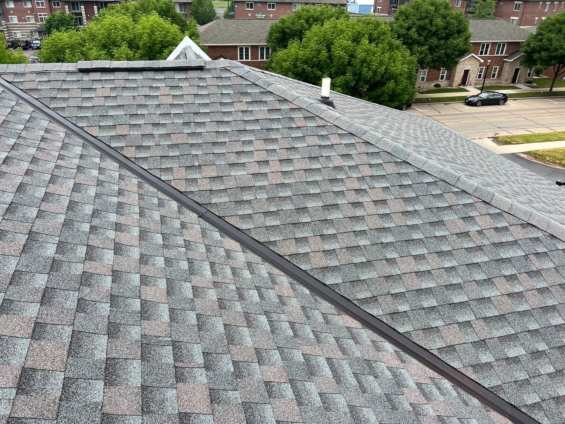 A high-angle view of a gray shingled roof with a diagonal ridge vent and a plumbing pipe, overlooking a parking lot.
