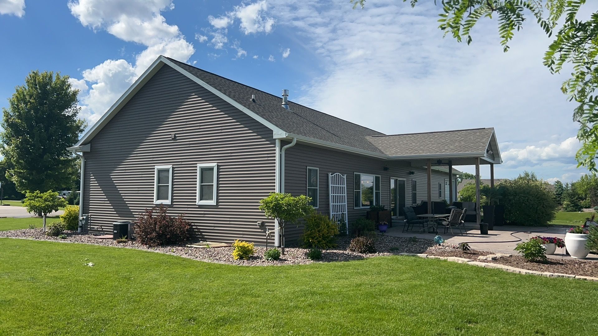 A gray-sided house with a shingled roof, a covered back patio, and a landscaped yard under a blue sky with white clouds.