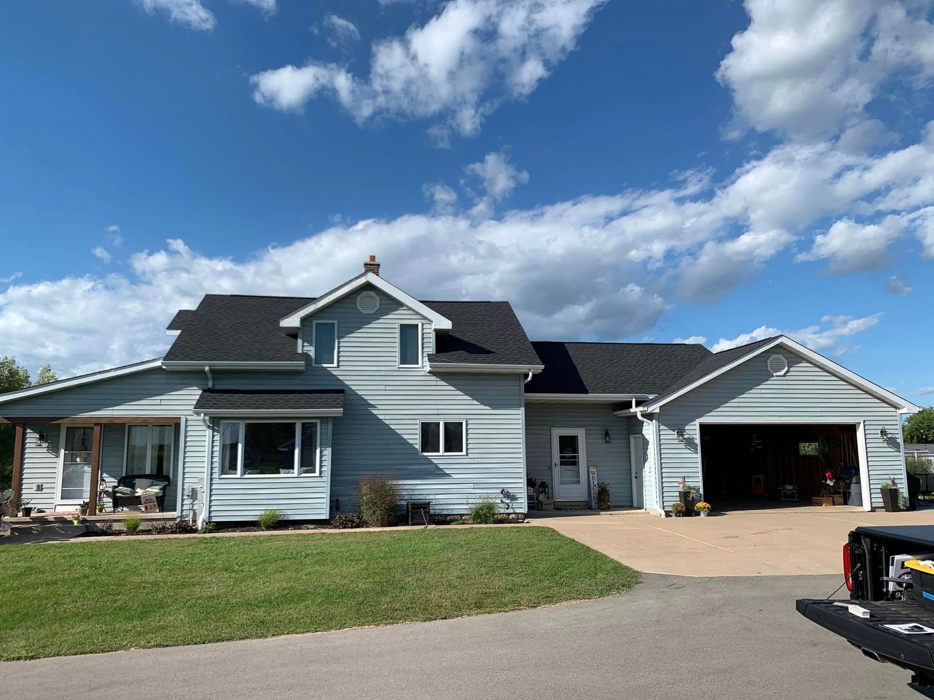 A light blue two-story house with a black roof and an attached garage, set against a bright blue sky with white clouds.