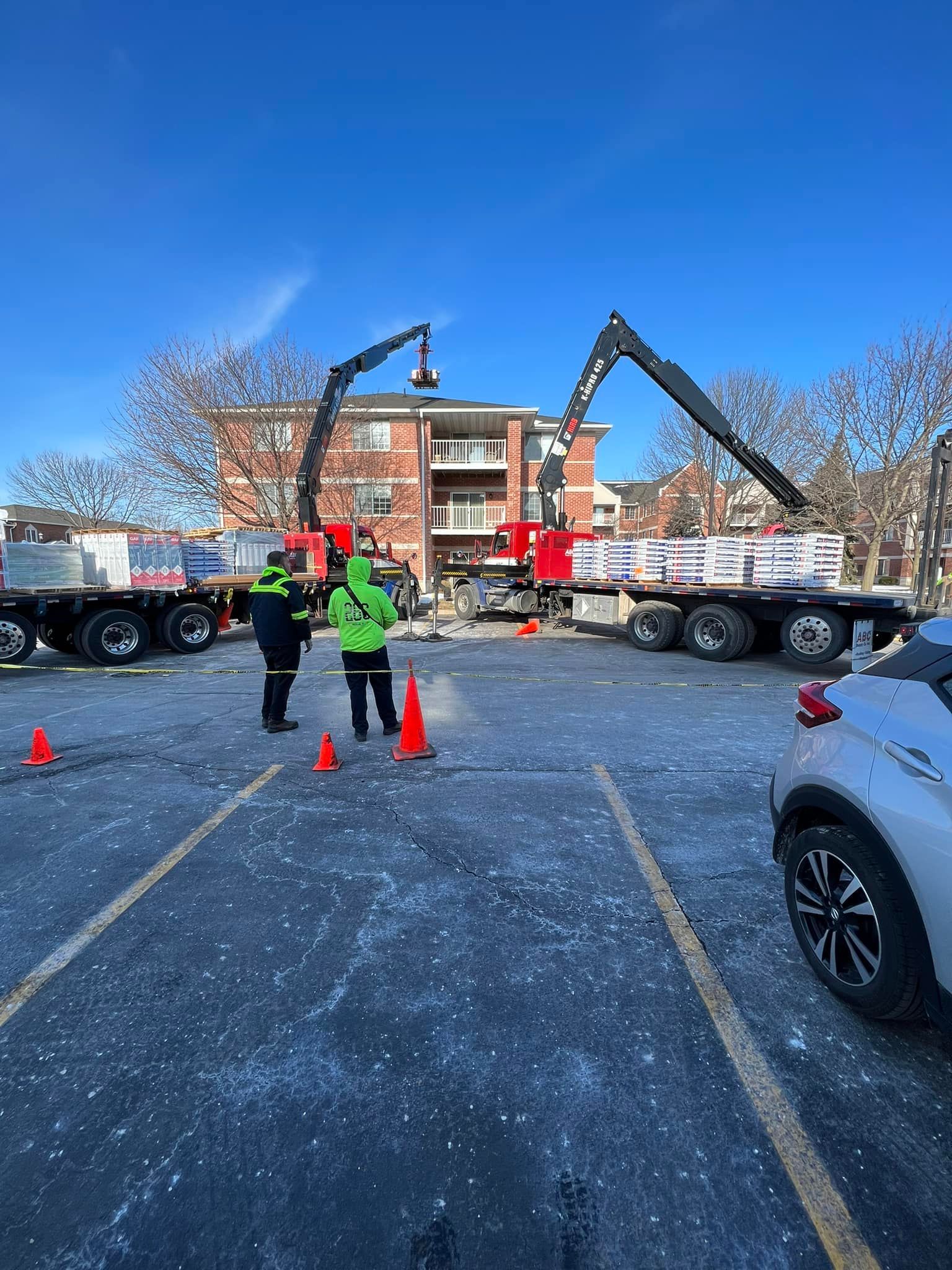 Two red boom trucks deliver building materials to a brick apartment building in a parking lot with two workers present.