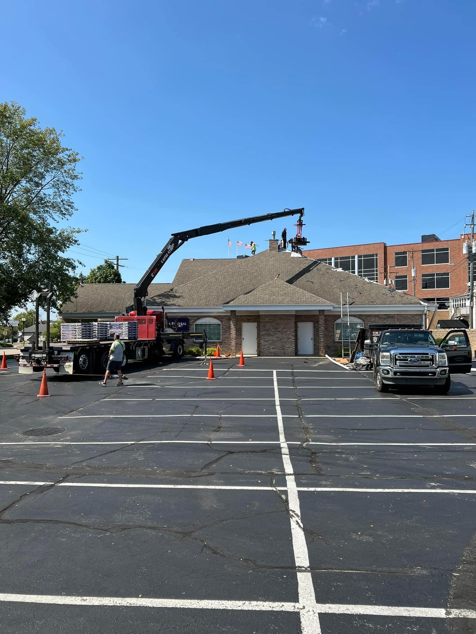 A crane truck parked in a parking lot lifts an object onto the roof of a brick building on a clear, sunny day.