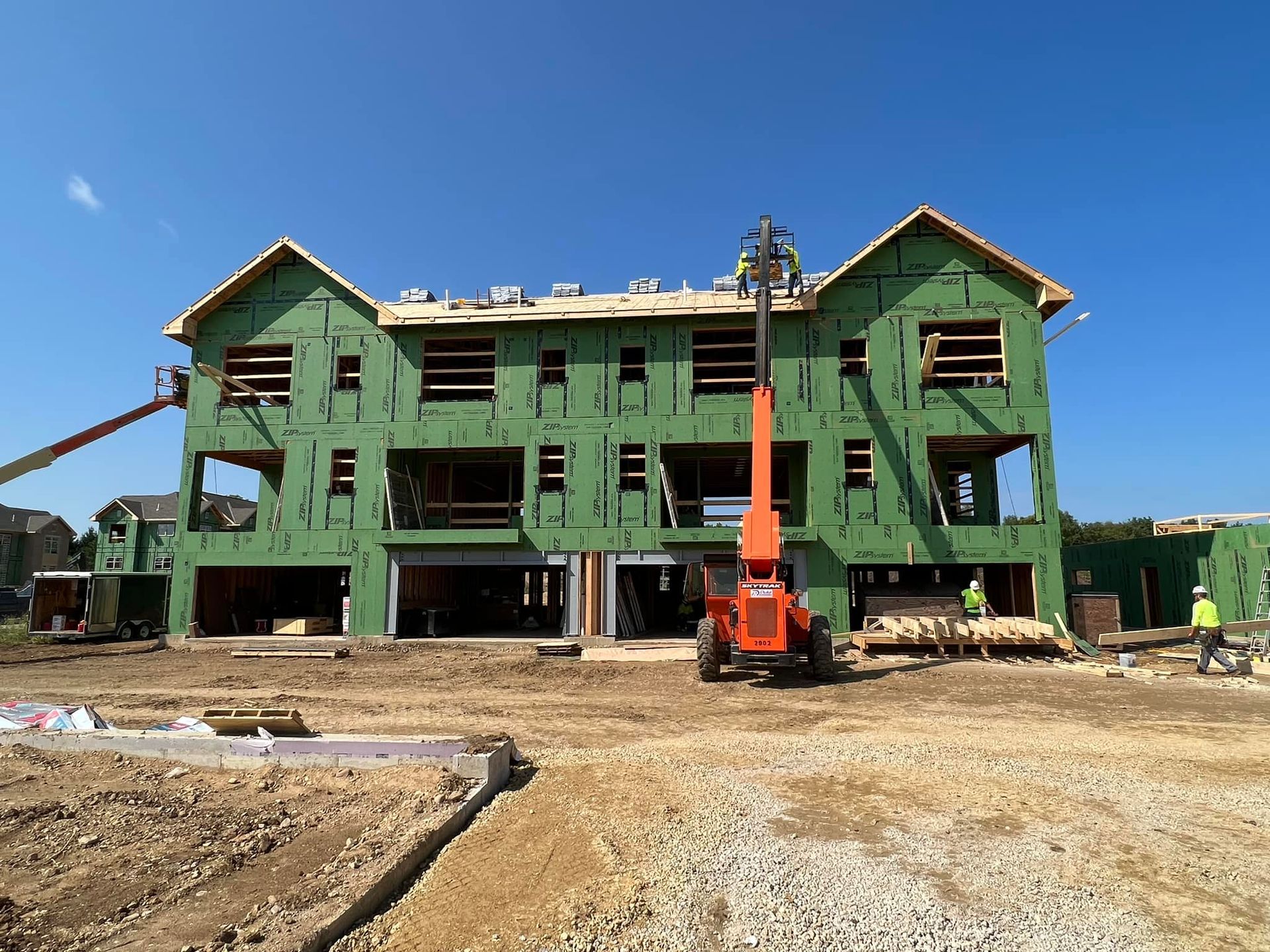 An orange telehandler lifts workers to the roof of a multi-story building under construction with green exterior sheathing.