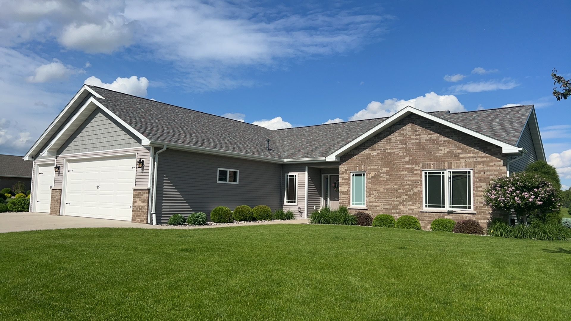 A tan brick and grey siding ranch-style house with a multi-car garage, set against a blue sky with white clouds.